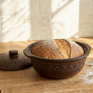 Könnte beinhalten: Ein brauner Keramik-Brotbacktopf mit einem Brot darin und der Deckel daneben. Der Topf hat dekorative Muster am Rand. Ein Anhänger mit der Aufschrift "CERAMIC BAKERY" ist befestigt.