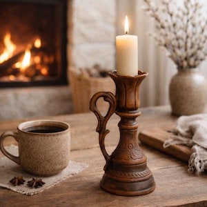 May include: A lit candle sits in a brown, ornate candlestick holder with a handle. A mug of coffee and star anise sit on a wooden table. A fireplace with a burning fire is in the background, creating a cozy atmosphere.