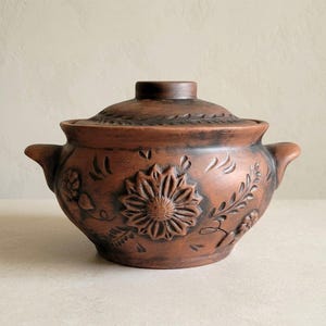 May include: A brown ceramic cooking pot with a lid. The pot features a large embossed sunflower design on the front, with floral and leaf patterns. It has two handles and a knob on the lid.