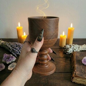 May include: A dark brown wooden chalice with a smooth, turned stem and a wide base. The cup features a visible wood grain pattern and a cylindrical shape. A faint, wispy golden vapor rises from the top rim of the vessel, suggesting warmth.