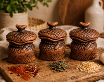 Puede incluir: Tres tarros de especias de cerámica marrón con tapa, con forma de sacos texturizados. Cada tarro tiene una etiqueta única: Paprika, Basil y Garlic. Los tarros están dispuestos sobre una tabla de madera, con montones de especias delante.