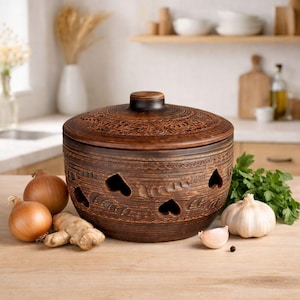 Könnte beinhalten: Ein brauner Knoblauchtopf aus Keramik mit Deckel, mit herzförmigen Aussparungen und dekorativen Schnitzereien. Der Topf ist von Zwiebeln, Knoblauch, Ingwer und Petersilie auf einer Holzoberfläche umgeben. Im Hintergrund ist eine Küchenszene zu sehen.