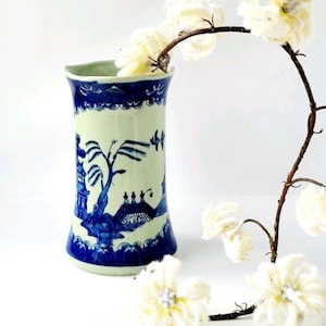 Puede incluir: Un jarrón de cerámica azul y blanco con un diseño tradicional de paisaje asiático. El jarrón tiene una forma acampanada y un patrón detallado de árboles, edificios y figuras. Una rama decorativa con flores blancas está en el fondo.