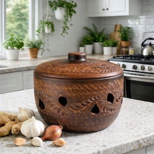 May include: A round, brown clay garlic keeper with a matching lid, featuring carved leaf patterns and teardrop-shaped ventilation holes around the base. The container is displayed alongside several whole garlic bulbs and ginger roots.