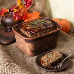Puede incluir: Una fuente rectangular de arcilla marrón que contiene un pastel de carne cocinado con hierbas por encima. Una rebanada de pastel de carne descansa sobre un pequeño plato de arcilla a juego con un tenedor de plata. La tapa está a un lado.