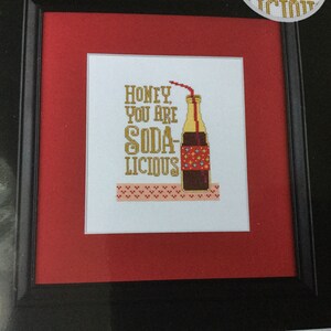 May include: Framed cross-stitch art with a black frame and red matting. The artwork features a soda bottle with a straw and the words "Honey, You Are Soda-licious" in gold lettering. A decorative border is at the bottom.