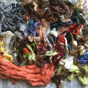 May include: A pile of colorful embroidery floss in various shades of orange, yellow, green, blue, brown, and gray. The floss is bundled and tangled, creating a textured and vibrant display. Ideal for embroidery and other craft projects.