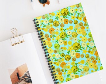 May include: A spiral-bound notebook with a vibrant floral pattern in yellow, orange, and green on a light blue background. The notebook is open on a white surface, with a gold paper clip and sunglasses visible.