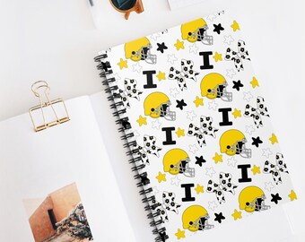 Op de afbeelding: Spiraalgebonden notitieboek met een witte omslag met een herhalend patroon van gele American footballhelmen, zwarte sterren, bloemenstrikken en de letter "I". Het notitieboek is open en toont blanco pagina's. Andere items liggen op het witte oppervlak.