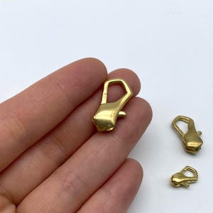Könnte beinhalten: Nahaufnahme von mehreren goldfarbenen Karabinerverschlüssen. Ein Verschluss wird in einer Hand gehalten, während zwei weitere in der Nähe positioniert sind. Die Verschlüsse haben einen rechteckigen Körper und eine gebogene Öffnung.