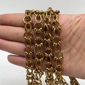 Puede incluir: Primer plano de una cadena dorada con un diseño intrincado. La cadena está compuesta por eslabones ovalados y en forma de lazo entrelazados, creando un patrón decorativo y elegante. La cadena se muestra sobre un fondo blanco.