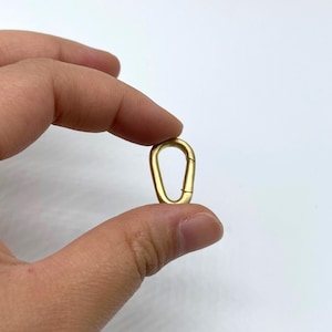 Pode incluir: Um pequeno mosquetão dourado. O mosquetão tem formato oval com um portão com mola. O mosquetão é segurado entre os dedos contra um fundo branco. O clipe tem aproximadamente 3,8 cm de comprimento.