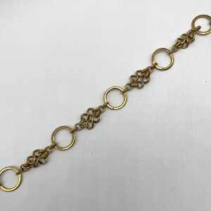 May include: A gold-colored chain necklace with a repeating pattern of open circles and decorative, intertwined shapes. The necklace is laid out on a white surface, showcasing its design and details. The chain is a jewelry item.