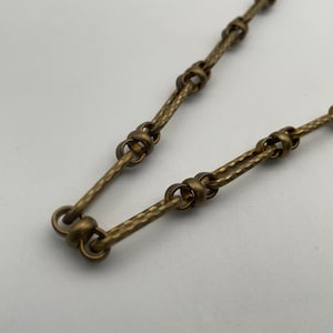 May include: Close-up of a vintage brass chain necklace. The chain features a textured, hammered design with connecting links and circular accents. The warm, antique brass color gives it a classic, timeless appeal.