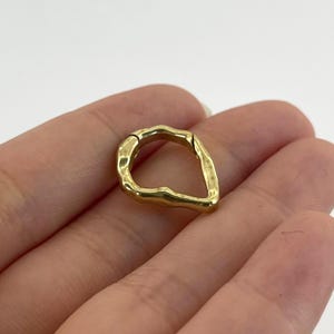 Pode incluir: Um anel de metal dourado com formato irregular. O anel tem uma superfície texturizada e um fecho articulado. O anel é segurado na mão de uma pessoa contra um fundo branco. É uma joia.
