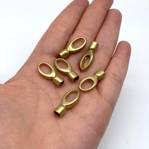 May include: Six small, gold-coloured oval-shaped metal findings. Each finding has a cylindrical base and an oval loop. The findings are displayed on a light background.