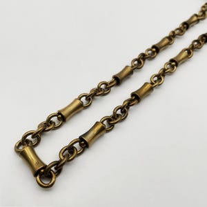 Puede incluir: Primer plano de un collar de cadena de latón vintage. La cadena presenta una serie de pequeños eslabones redondeados intercalados con segmentos cilíndricos en forma de bambú. El metal tiene un acabado cálido y antiguo.
