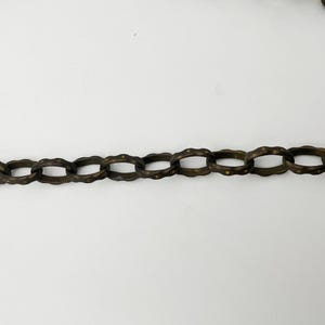 Puede incluir: Primer plano de una sección de una cadena de color bronce. Los eslabones de la cadena tienen forma ovalada con un diseño texturizado y ondulado. El fondo es blanco liso, resaltando los detalles y el color de la cadena.