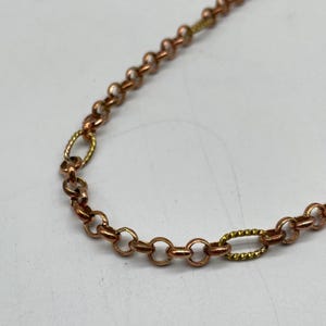 Puede incluir: Primer plano de un collar de cadena de color cobre con eslabones ovalados y circulares. La cadena tiene secciones de eslabones texturizados de color dorado. El collar está sobre un fondo blanco.