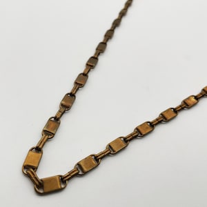 Puede incluir: Primer plano de un collar de color bronce. La cadena está compuesta por eslabones rectangulares conectados por eslabones más pequeños y redondeados. El metal tiene un aspecto ligeramente envejecido, con variaciones de color. El collar se muestra sobre un fondo blanco liso.