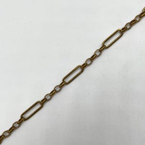 May include: Close-up of a gold-colored chain with a repeating pattern of rectangular and circular links. The chain is set against a white background, and the links are connected in a linear fashion.
