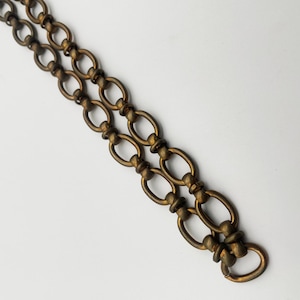 Puede incluir: Primer plano de una cadena de latón vintage con eslabones ovalados. La cadena está compuesta por dos hilos paralelos, cada uno con eslabones entrelazados. El metal tiene una pátina cálida y envejecida, lo que sugiere que es una pieza antigua o vintage. La cadena está sobre un fondo blanco.