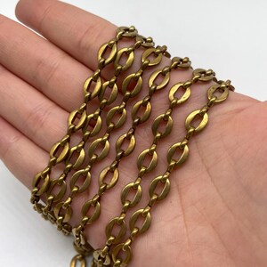 Könnte beinhalten: Nahaufnahme einer Vintage-Halskette mit goldfarbener Kette. Die Kette hat ein ovales Kettenglied-Design, wobei jedes Glied durch ein kleineres, längliches Glied verbunden ist. Die Halskette wird vor einem neutralen Hintergrund gezeigt.