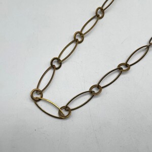 May include: A gold-coloured chain necklace with a series of elongated oval links connected by small circular rings. The chain is displayed against a white background, highlighting its metallic sheen and simple design. This necklace is suitable for everyday wear.