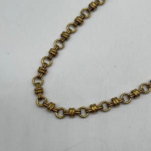 May include: Close-up of a gold-coloured chain necklace. The chain features small, round links connected by cylindrical bars. The necklace is set against a white background.