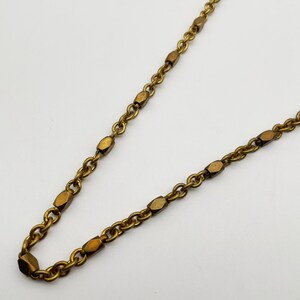 Op de afbeelding: Close-up van een goudkleurige ketting met een delicaat kettingontwerp. De ketting heeft kleine, ovaalvormige schakels afgewisseld met grotere, geometrisch gevormde segmenten. De ketting staat tegen een witte achtergrond.