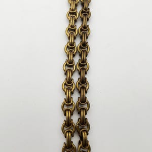 Op de afbeelding: Close-up van twee parallelle antiek messingkleurige kettingen. Elke ketting is samengesteld uit in elkaar grijpende cirkelvormige schakels, waardoor een gestructureerd, decoratief patroon ontstaat. De kettingen staan tegen een effen witte achtergrond.