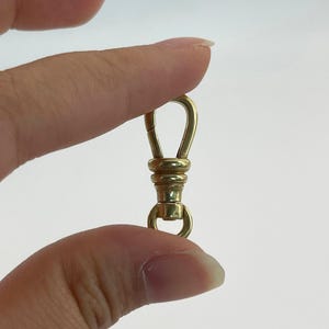 May include: A small, gold-coloured swivel clasp with a spring-loaded hook and a ring at the bottom. The clasp is held between fingers against a white background. The metal has a polished finish.