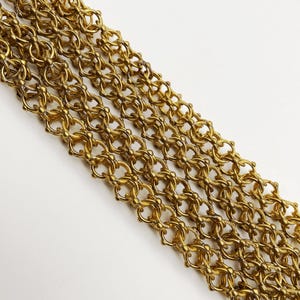 Puede incluir: Primer plano de un accesorio dorado estilo cota de malla. El intrincado diseño presenta elementos circulares y florales interconectados. La cota de malla está dispuesta diagonalmente sobre un fondo blanco, mostrando su textura y detalle.