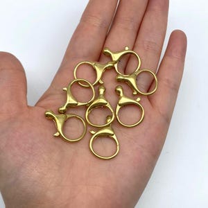 May include: A collection of small, gold-colored lobster claw clasps. Each clasp features a circular ring and a lever-activated closure mechanism. The clasps are displayed against a light background, showcasing their metallic sheen and design.