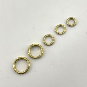 Può includere: Una collezione di anelli a molla rotondi color oro di varie dimensioni. Le chiusure sono disposte diagonalmente, mostrando il loro design circolare e la finitura metallica. Queste chiusure sono adatte per la creazione di gioielli o progetti artigianali.