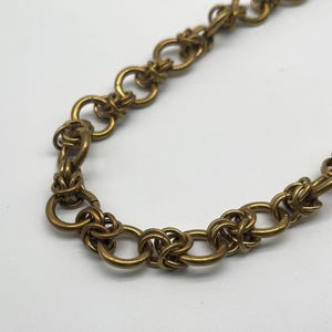 May include: Close-up of a gold-toned chain necklace. The chain features a series of interconnected rings and woven knot-like details. The metal has a slightly aged appearance, suggesting a vintage or antique style. The necklace is displayed against a white background.