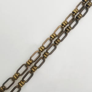 May include: Two parallel antique brass chains with oval and cylindrical links. The chains are set against a white background, creating a contrast that highlights the intricate design and aged finish of the metal.