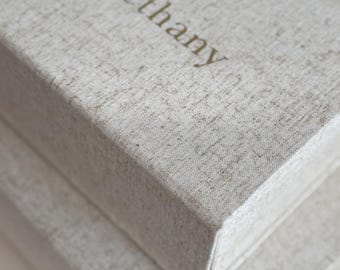 Może przedstawiać: Zbliżenie dwóch prostokątnych pudełek ułożonych jedno na drugim. Pudełka pokryte są jasnobeżową tkaniną o teksturowanym wyglądzie. Słowo "Bethany" jest wydrukowane złotymi literami na górnym pudełku.