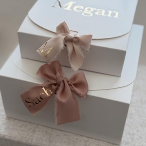 May include: Two white gift boxes, one stacked on top of the other. The top box has the name "Megan" in gold lettering. Each box has a pink ribbon with gold lettering. The bottom ribbon says "Sach".