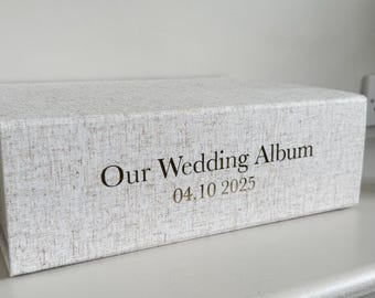 Może przedstawiać: Prostokątny album na zdjęcia z jasnoszarą, teksturowaną okładką z tkaniny. Z przodu nadrukowano czarny napis "Our Wedding Album" oraz datę "04.10.2025" wyraźną, ciemną czcionką szeryfową, wyśrodkowaną na bocznej części okładki.