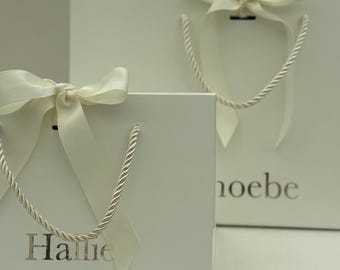 Może przedstawiać: Dwie białe torby na prezenty z kremowymi kokardkami i sznurkowymi uchwytami. Torby mają złote napisy „Hallie” i „Phoebe”. Odpowiednie dla sklepu z prezentami lub butiku.