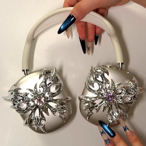 May include: Silver headphones with ornate, metallic floral designs on the earcups. The headphones have a white headband and are accented with a pink and purple gem. The earcups have a unique, stylized, and decorative design.