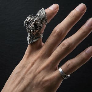 May include: A detailed silver ring featuring a dragon-like design with sharp teeth and textured scales. The ring is worn on a finger, with a smaller, plain silver band on an adjacent finger. The background is dark.