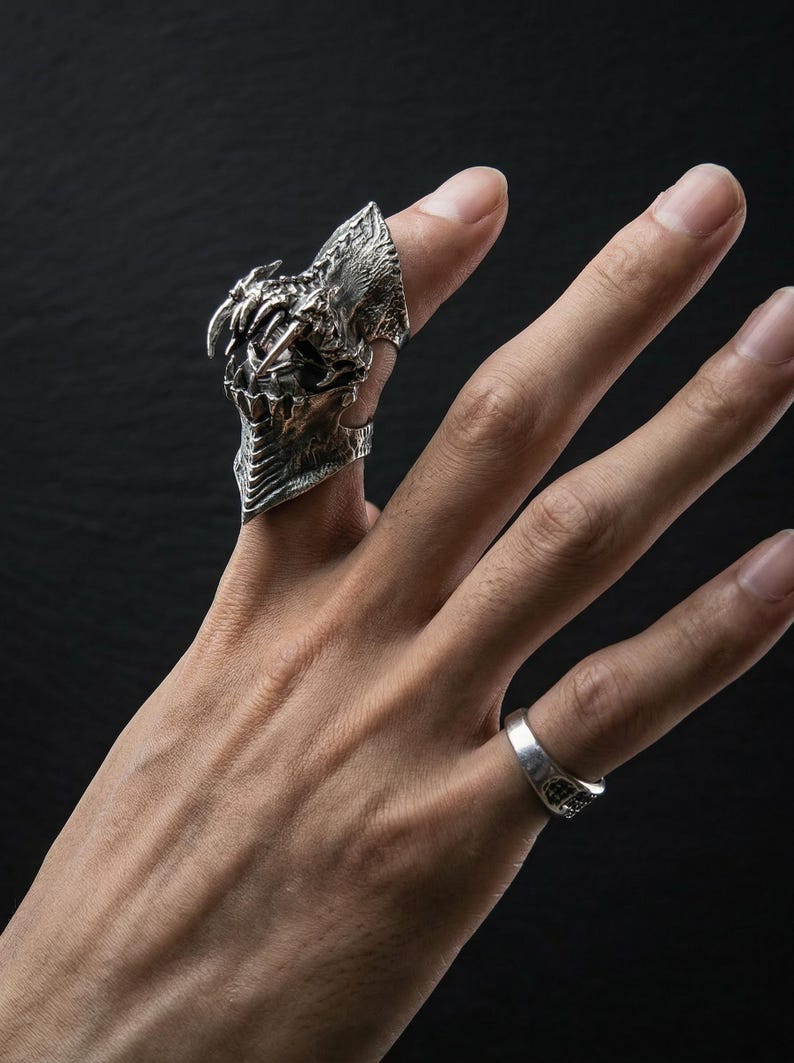 May include: A detailed silver ring featuring a dragon-like design with sharp teeth and textured scales. The ring is worn on a finger, with a smaller, plain silver band on an adjacent finger. The background is dark.