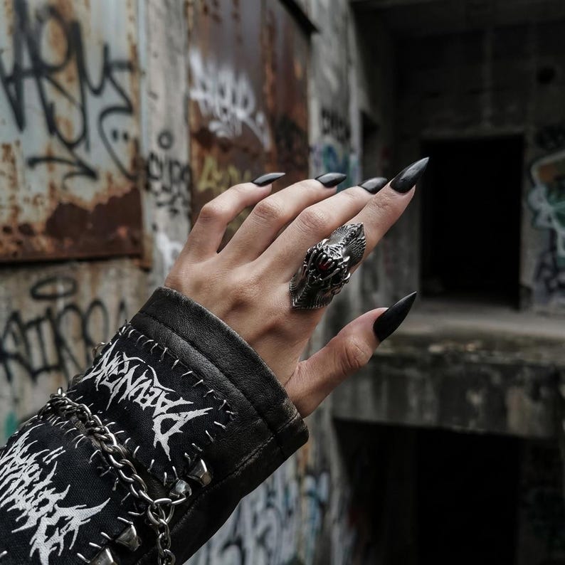 May include: A silver-toned ring with a dragon-like design, featuring a red stone accent. The ring is worn on a hand with long, black fingernails. The person is wearing a black leather jacket with white text and silver chain details.