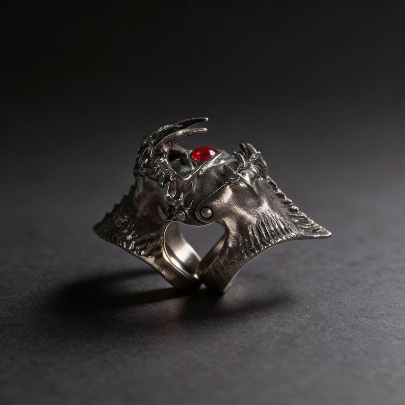 May include: A silver-toned ring with an ornate, textured design. The ring features a prominent red gemstone at the center and intricate detailing, suggesting a fantasy or gothic theme. The ring's design includes horns and a crown-like structure.