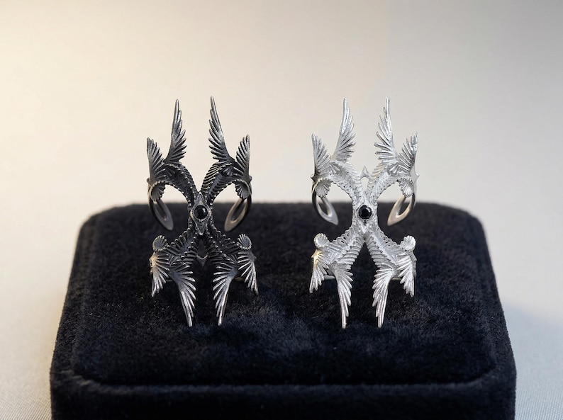 Puede incluir: Dos anillos ornamentados, uno negro y otro plateado, se exhiben sobre una superficie de terciopelo negro. Cada anillo presenta un dise&ntilde;o complejo y puntiagudo con una piedra oscura central. Los anillos tienen una est&eacute;tica art&iacute;stica &uacute;nica.