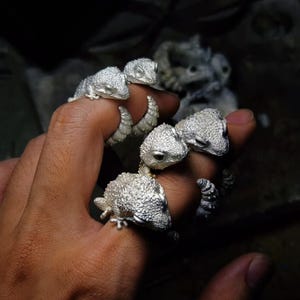May include: Three silver-toned rings, each featuring a detailed, textured lizard design that wraps around the finger. The metallic surfaces have a granular, matte finish, with the lizard heads positioned prominently on the outer side of each ring.