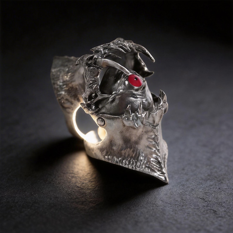 May include: A detailed silver ring with a dragon-like design. The ring features a red gemstone eye and intricate detailing, with a textured finish. The ring has a unique, open design, and is set against a dark background.