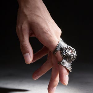 May include: A hand wearing two silver rings. One ring features a detailed dragon design, while the other has a simpler, textured band. The rings are set against a dark background, highlighting their intricate details and metallic sheen.
